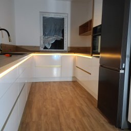 Mebstar - Nowoczesna kuchnia z białymi szafkami, drewnianym blatem i oświetleniem LED pod szafkami. Widoczny czarny, duży lodówka. Okno zakryte folią ochronną. Podłoga drewnopodobna.