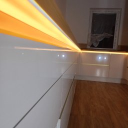 Mebstar - Nowoczesna, biała kuchnia z oświetleniem LED podszafkowym, widok z dołu, parkiet na podłodze, okno w tle. Minimalistyczny design i ciepłe światło.