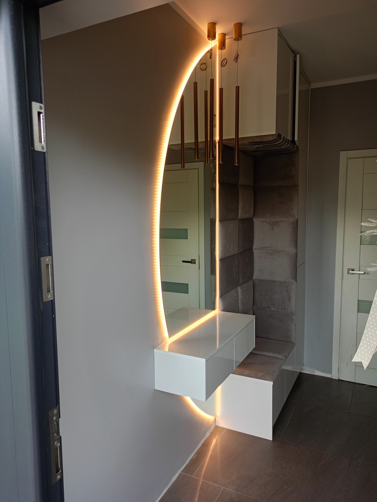Nowoczesna zabudowa przedpokoju z lustrem podświetlanym LED, tapicerowanym panelem ściennym i wiszącą szafką z oświetleniem punktowym. Minimalistyczny design, szaro-biała kolorystyka.
