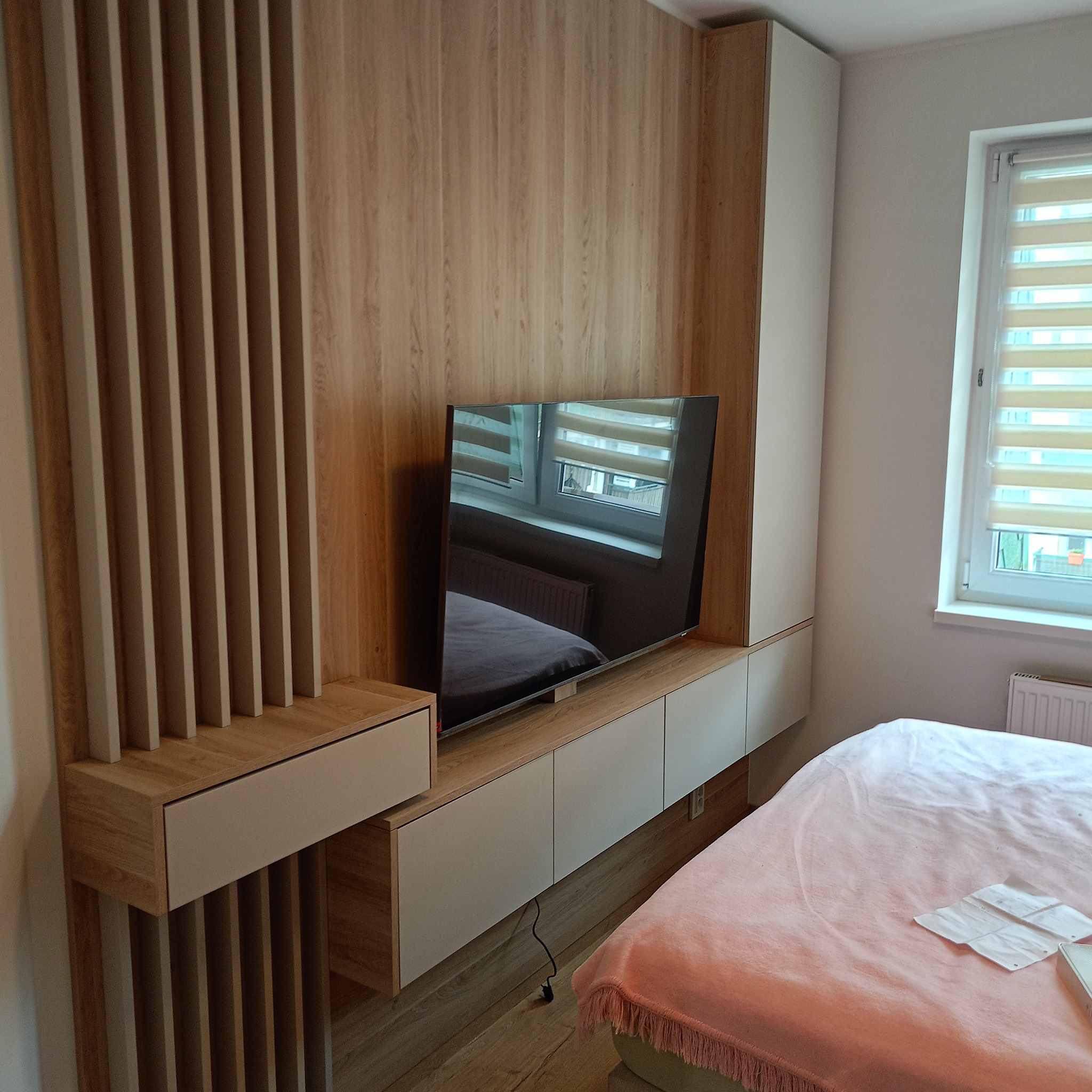 Nowoczesna szafa na wymiar z drewnianymi lamelami i białymi frontami, wkomponowana w aranżację sypialni z telewizorem. Widoczne detale wykończenia i oświetlenie.