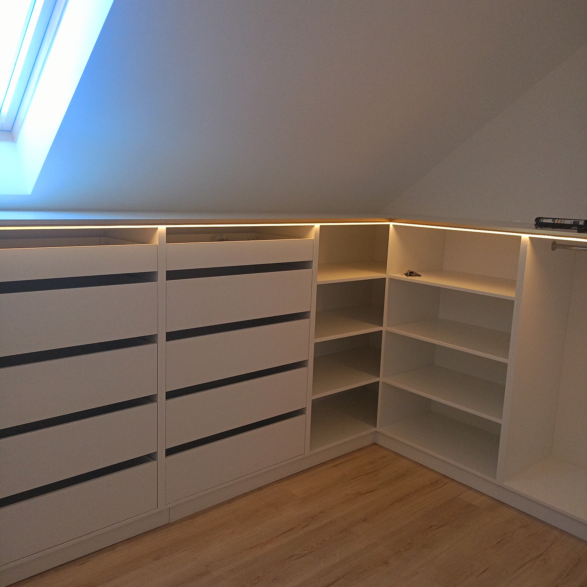 Biała szafa na poddaszu ze szufladami i półkami, oświetlona taśmą LED, drewniana podłoga, okno dachowe. Zabudowa pod skosem.