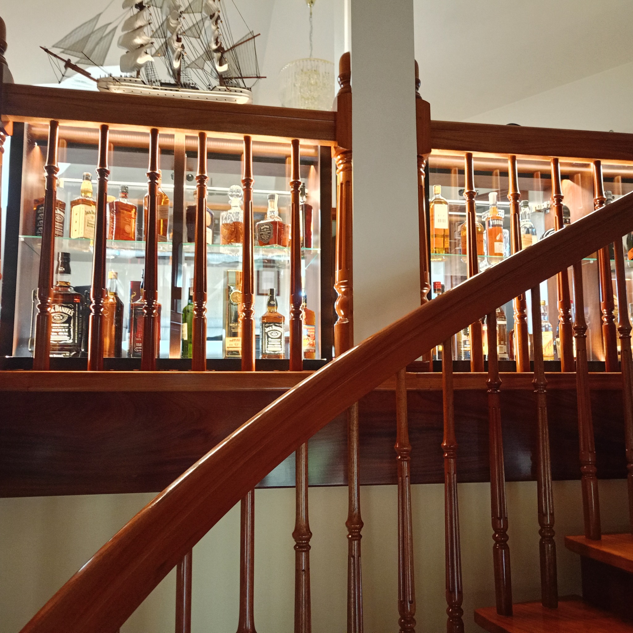 Drewniana balustrada schodów z dekoracyjną szafką podświetlaną LED, wypełnioną butelkami alkoholu i modelem żaglowca na górze. Ciepłe oświetlenie.