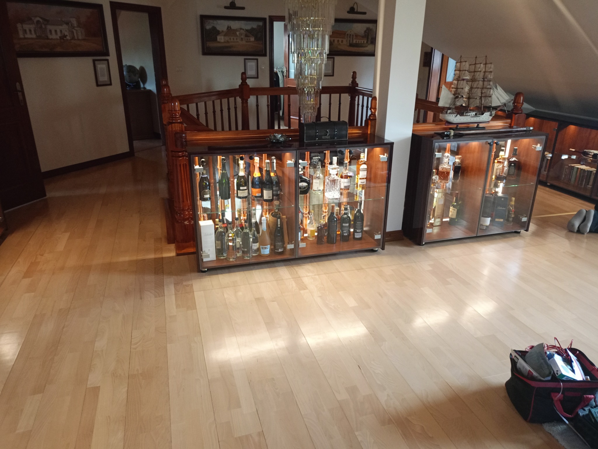 Eleganckie, przeszklone barki z oświetleniem, eksponujące kolekcję alkoholi w luksusowym wnętrzu z drewnianą podłogą i schodami. Na górze dekoracyjny żyrandol i model żaglowca.