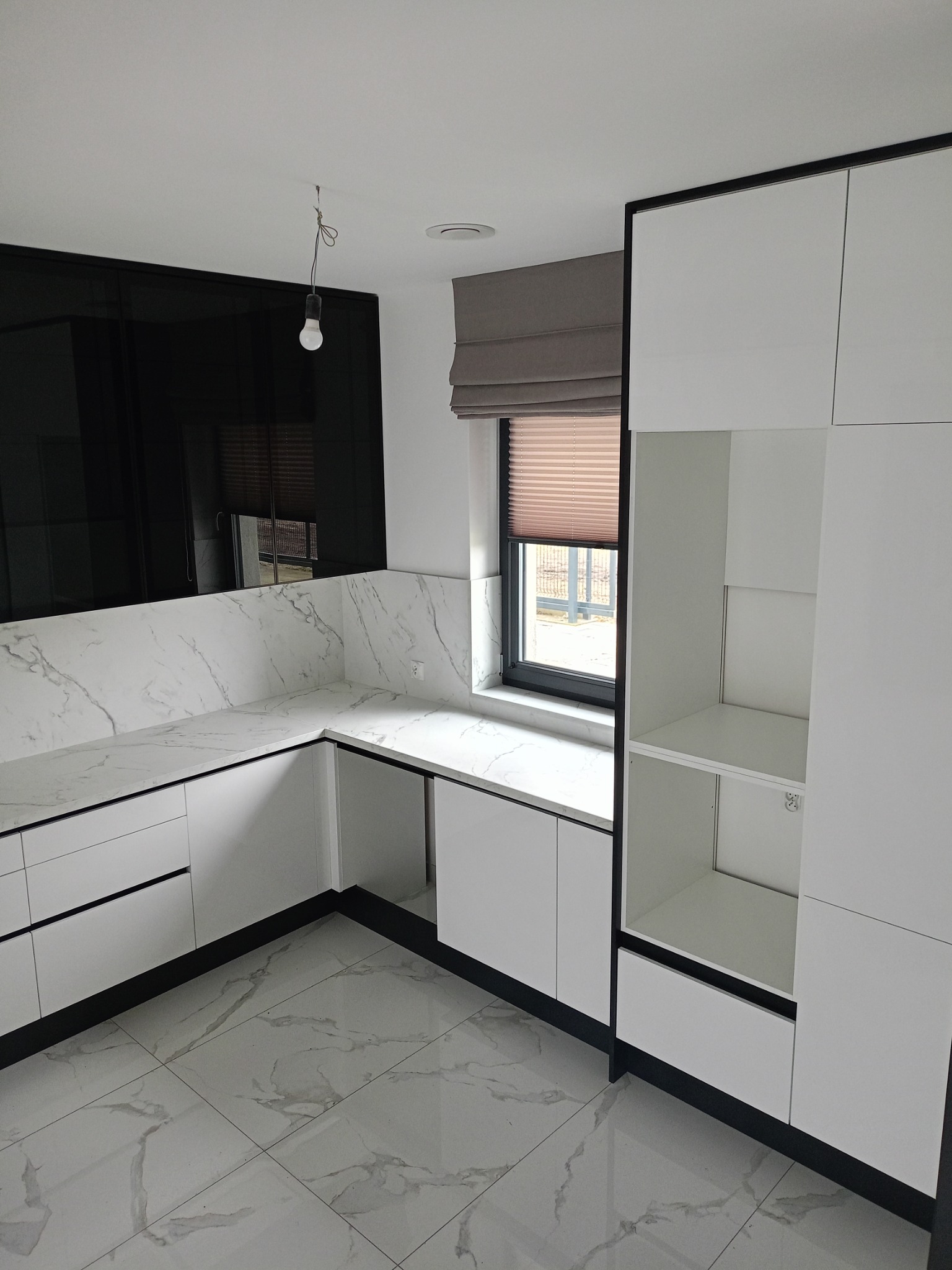 Nowoczesna kuchnia z białymi szafkami, blatem imitującym marmur i czarnymi akcentami. Widoczne okno z roletą i minimalistyczna lampa. Podłoga z płytek imitujących marmur.