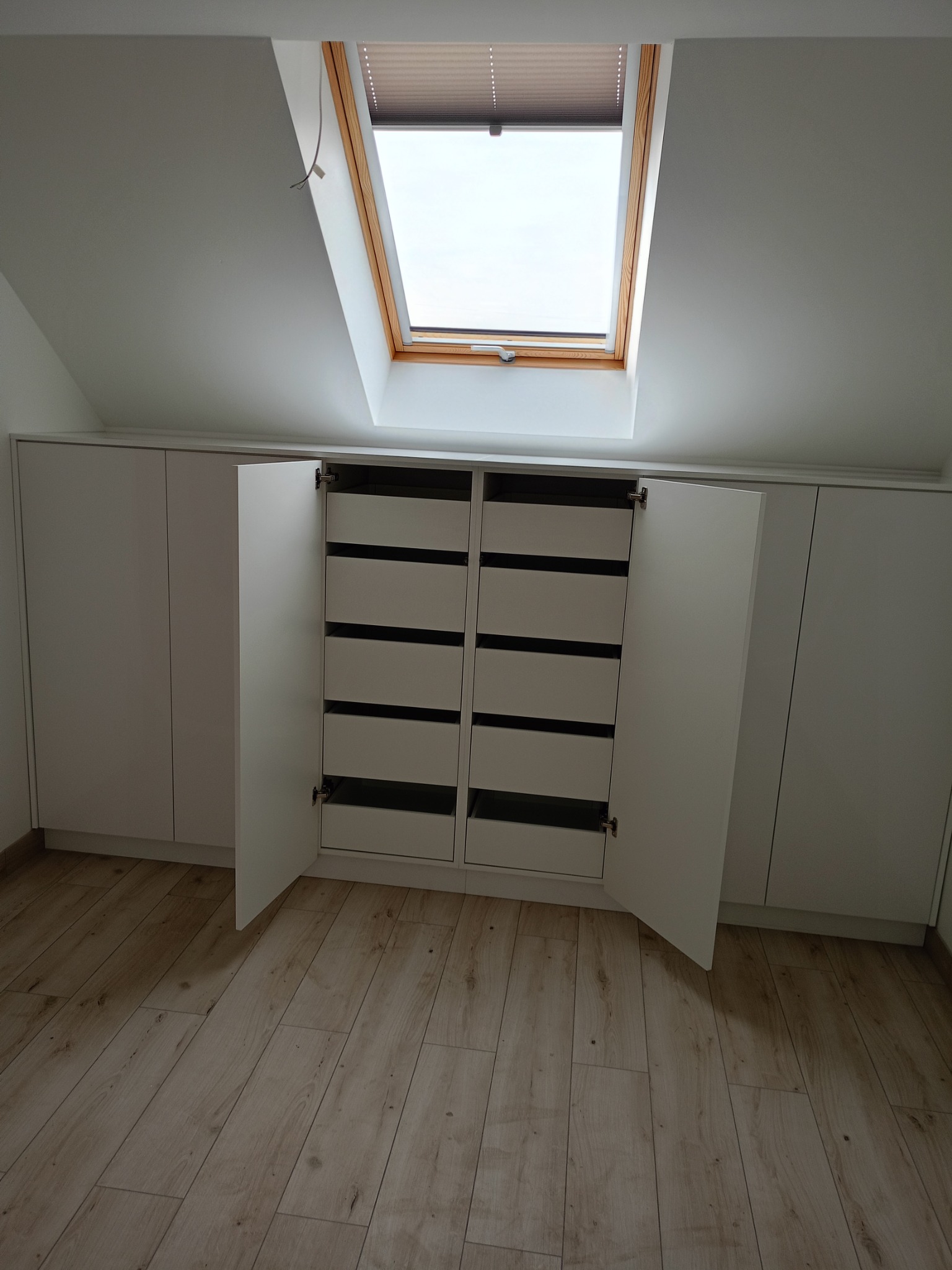 Biała szafa na poddaszu ze skośnym oknem dachowym. Otwarta szafa z szufladami, jasna podłoga. Minimalistyczny design, zabudowa pod skosem.