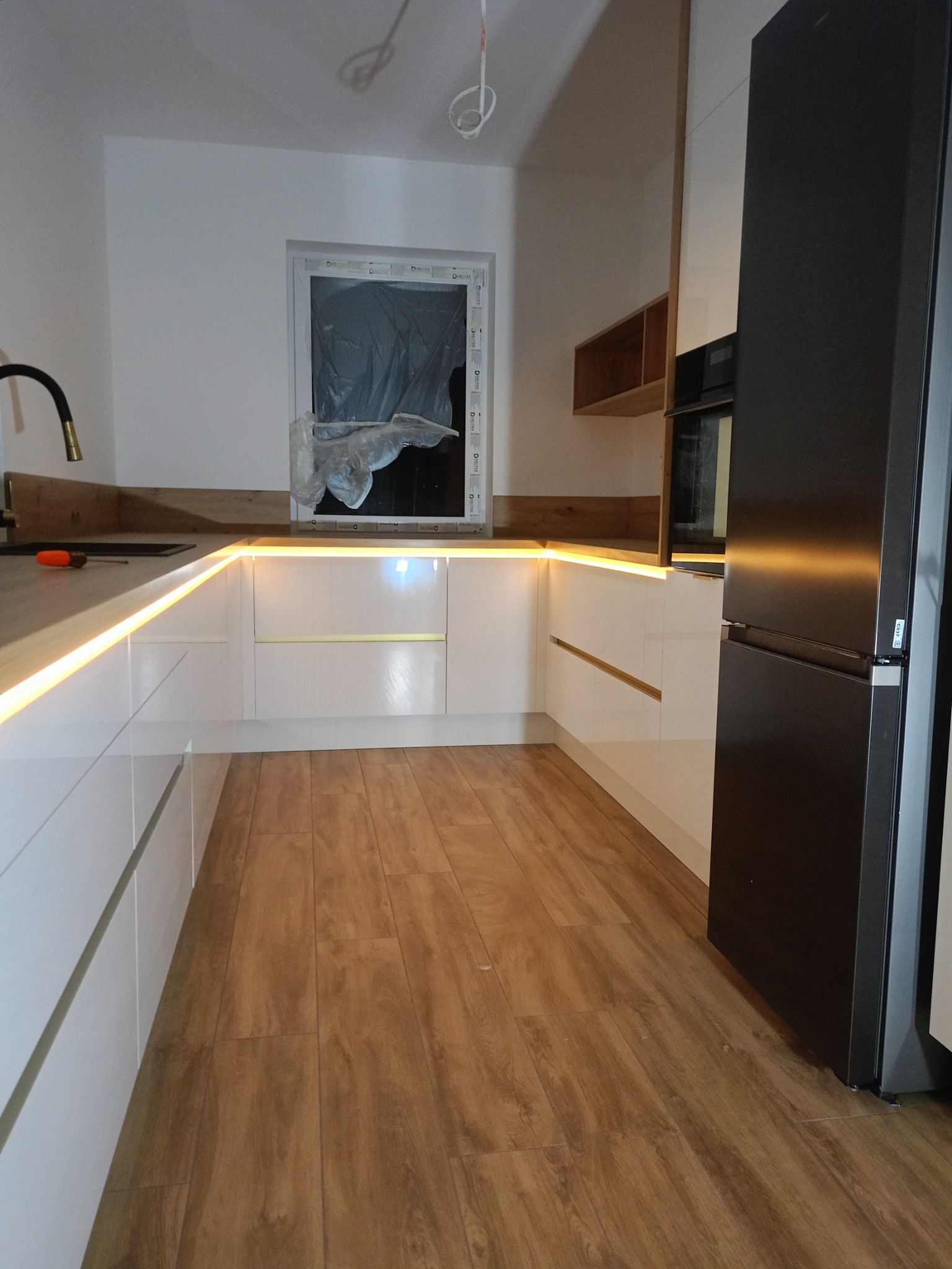 Nowoczesna kuchnia z białymi szafkami, drewnianym blatem i oświetleniem LED pod szafkami. Widoczny czarny, duży lodówka. Okno zakryte folią ochronną. Podłoga drewnopodobna.