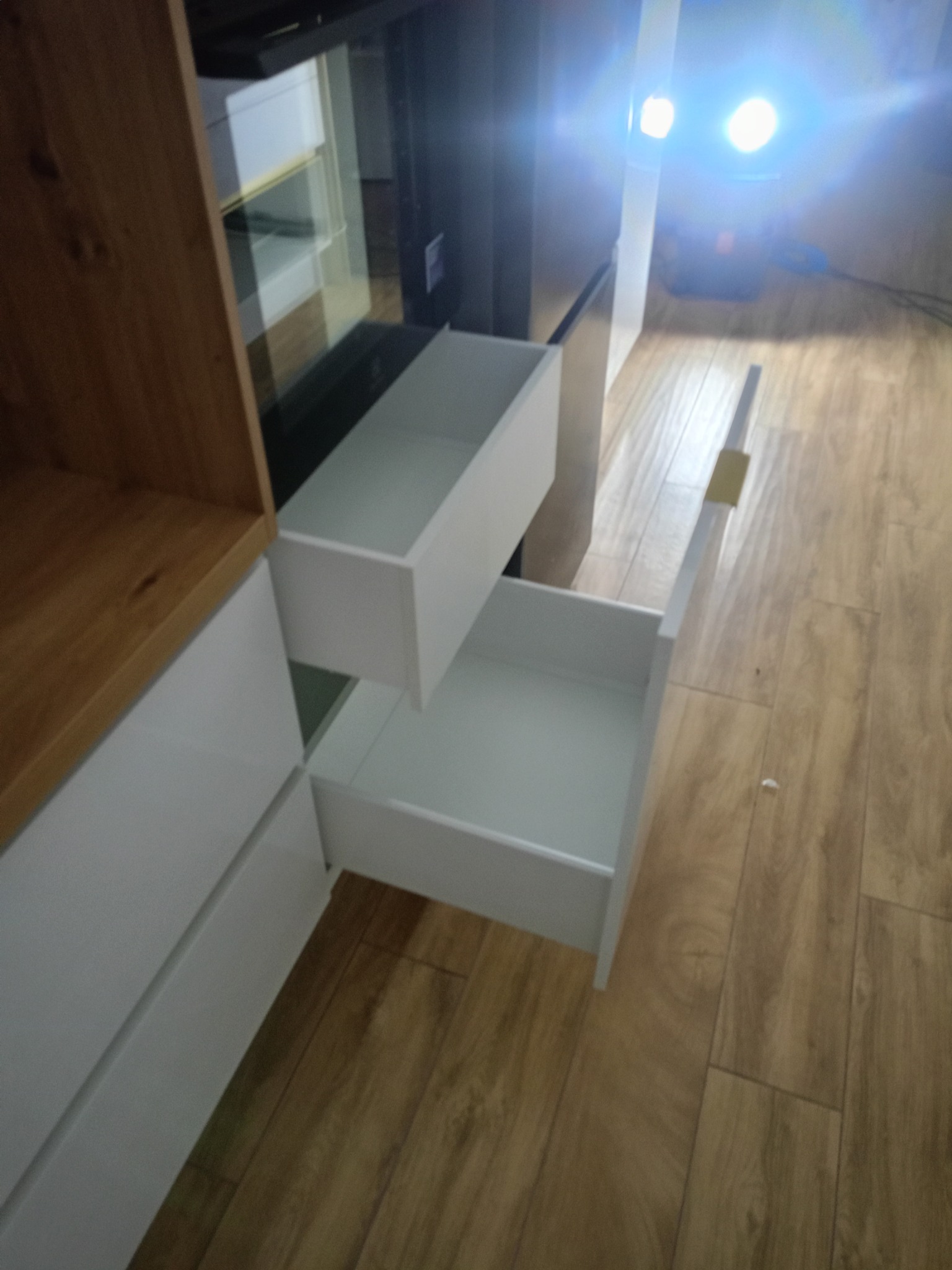 Wysuwane białe szuflady w szafce z drewnianym blatem, widok z dołu pod kątem, ukazujący mechanizm szuflady i drewnianą podłogę. Część szafki jest poza kadrem.