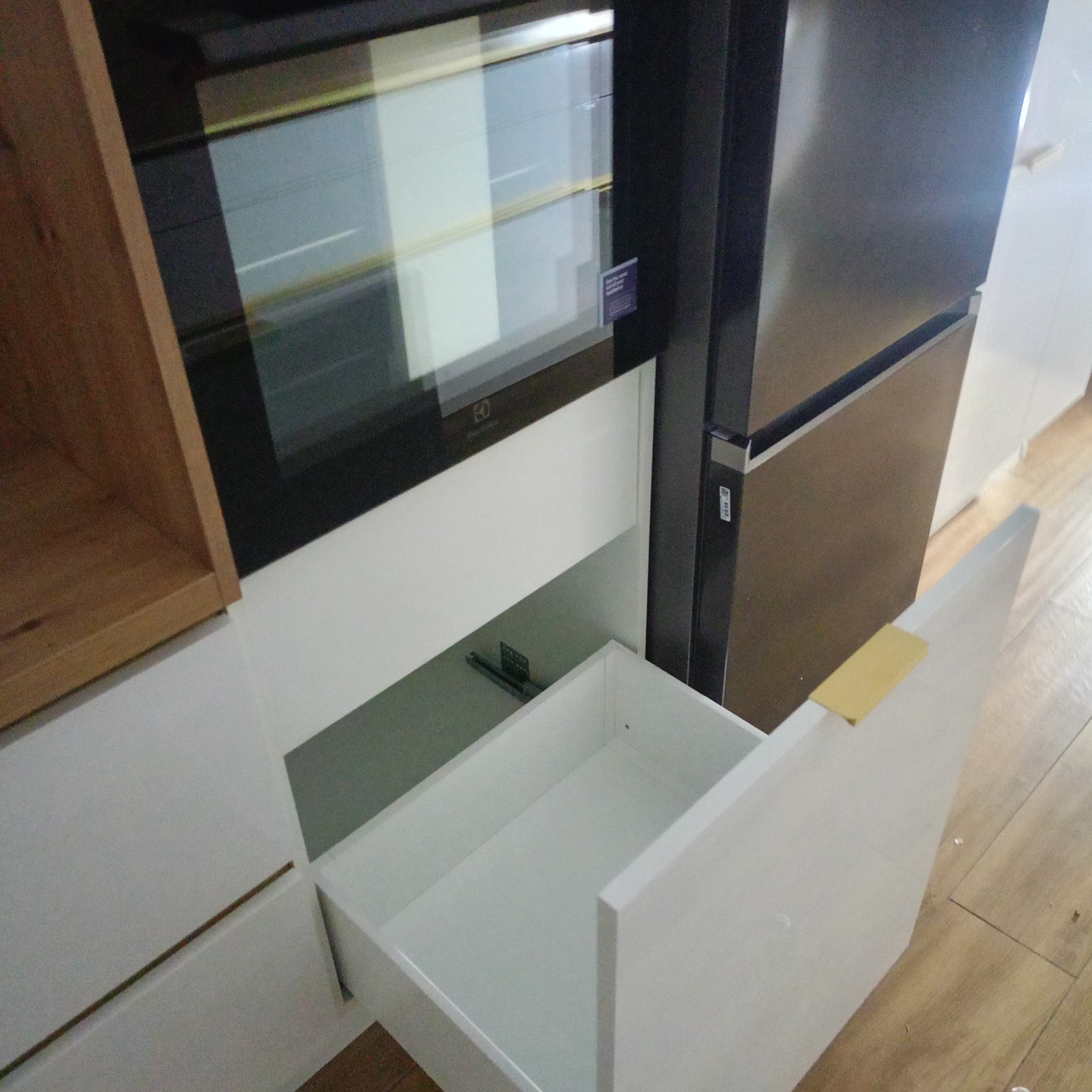 Otwarta, biała szuflada w zabudowie kuchennej z piekarnikiem i lodówką. Widoczne drewniane elementy wykończenia i podłoga. Minimalistyczny design kuchni.