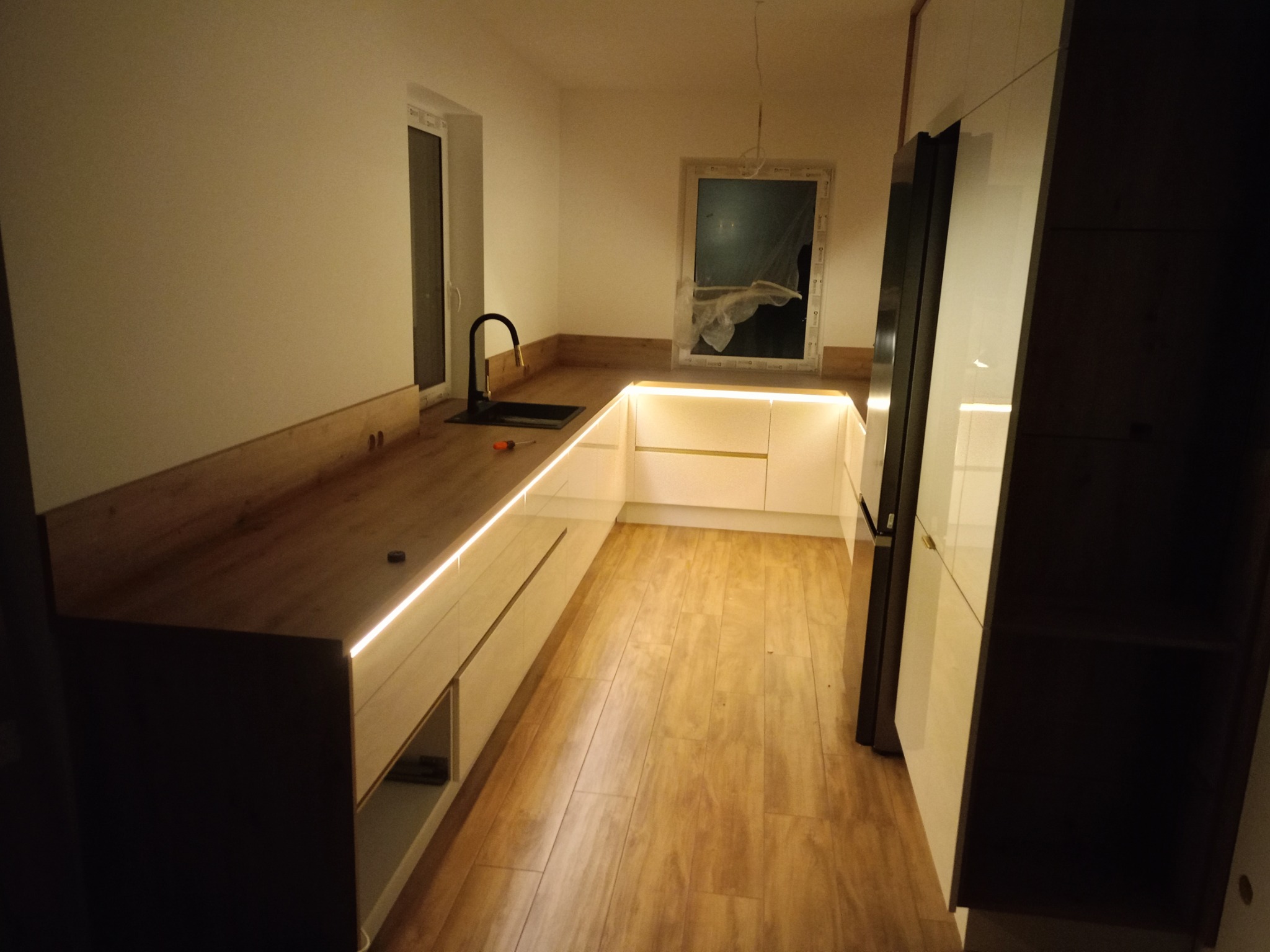 Nowoczesna kuchnia na wymiar w Stargardzie: białe fronty, drewniany blat z oświetleniem LED, czarna armatura i lodówka w zabudowie.