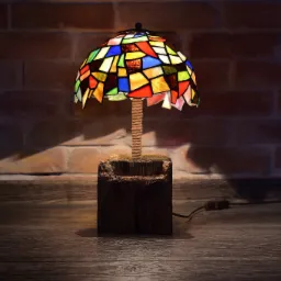 Lampa witrażowa w stylu Tiffany z kolorowymi szkiełkami, osadzona na drewnianej podstawie z jutowym oplotem, na tle ceglanej ściany i drewnianej podłogi.