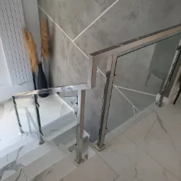 Szklana balustrada schodowa z nierdzewnymi słupkami, marmurowe schody i podłoga, szara ściana z geometrycznym wzorem, dekoracyjne trawy pampasowe w czarnym wazonie.