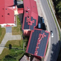 Widok z lotu ptaka na budynki z czerwonymi dachami, na których zamontowano panele fotowoltaiczne; obok droga z jadącym samochodem i strumień otoczony drzewami.