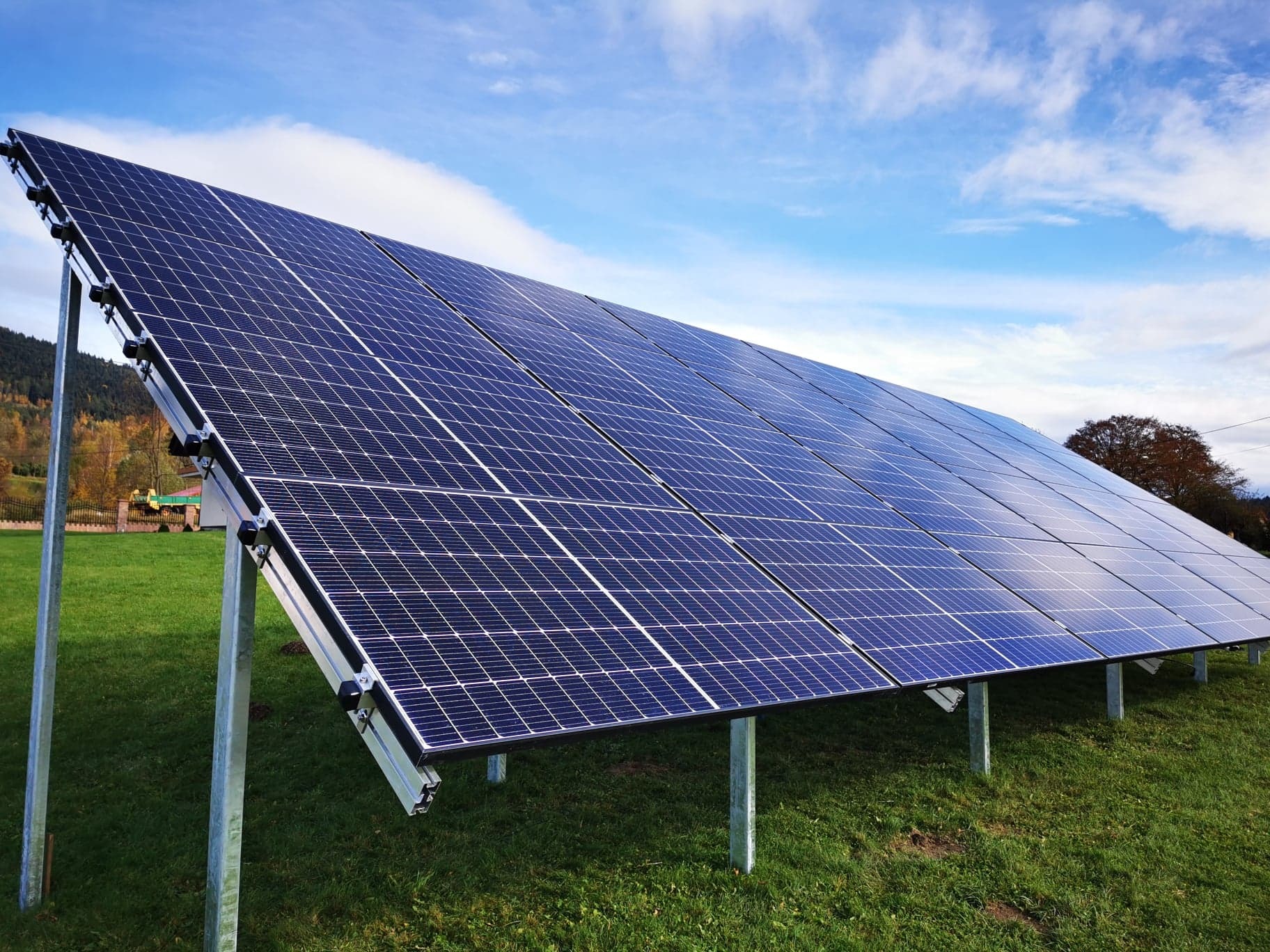 Panel słoneczny zamontowany na metalowych wspornikach na trawiastym terenie, błękitne niebo z chmurami w tle, widok na oddalony las i zabudowania.