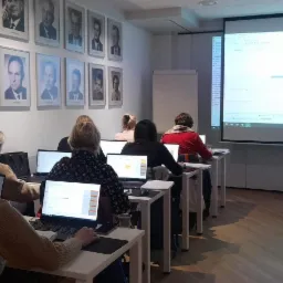 Szkolenie z zakresu kadr: Grupa kobiet siedzi przy biurkach z laptopami, na ekranie projektora widoczny arkusz kalkulacyjny z zaznaczonymi datami, na ścianie portrety w ramach.