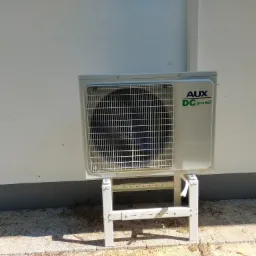 Zewnętrzna jednostka klimatyzacji AUX DC Inverter zamontowana na metalowym stelażu przy białej ścianie budynku.