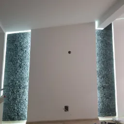 Ściana z dekoracyjną okładziną z kamienia naturalnego, podświetlona pionowymi pasami LED, gniazdko elektryczne na białej ścianie, widoczne narzędzia i materiały budowlane na podłodze.