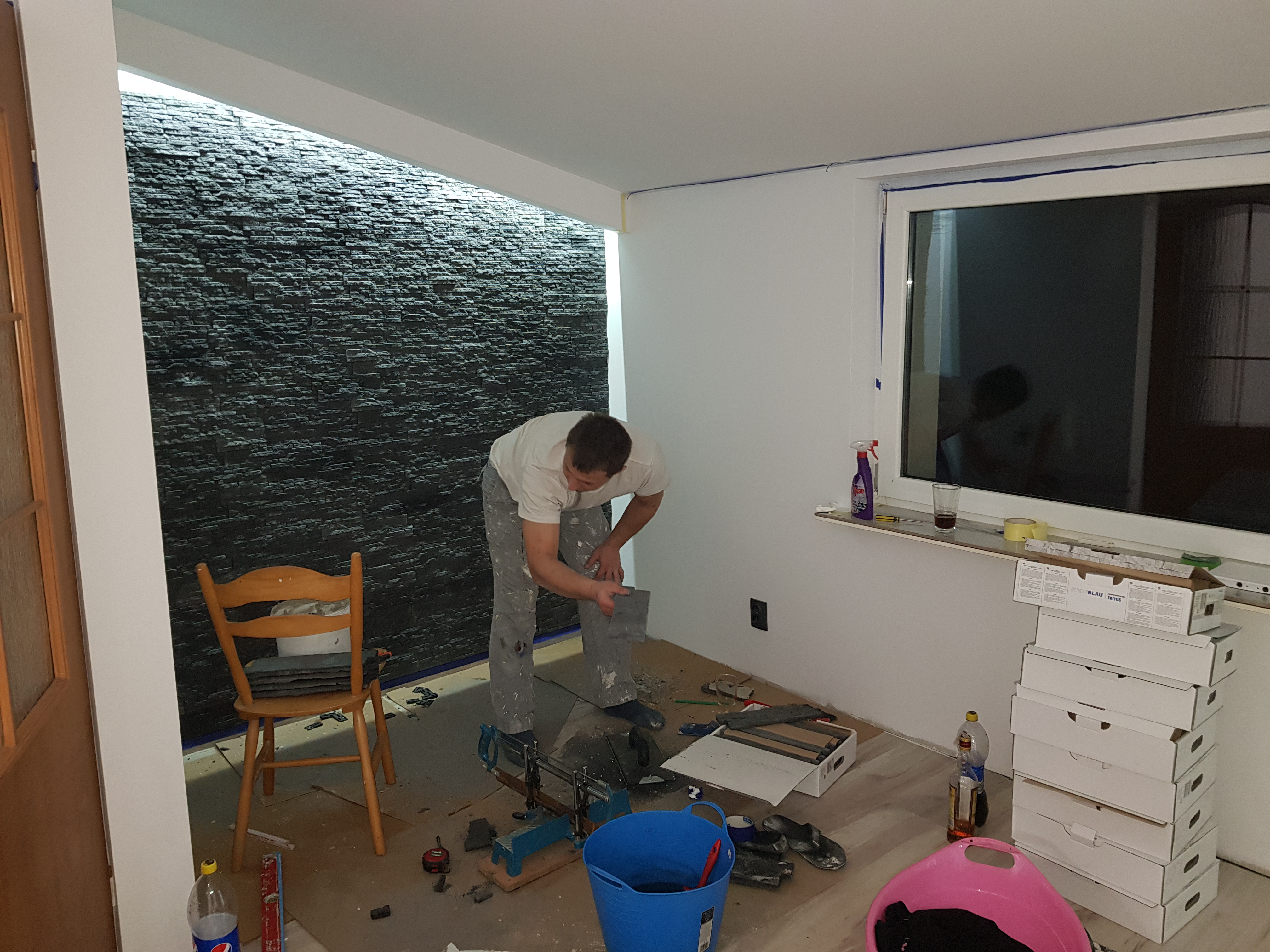 Pracownik w trakcie układania kamiennej okładziny na ścianie w pomieszczeniu, widoczne narzędzia, pocięte płytki, stos pudełek po okładzinie i drewniane krzesło.