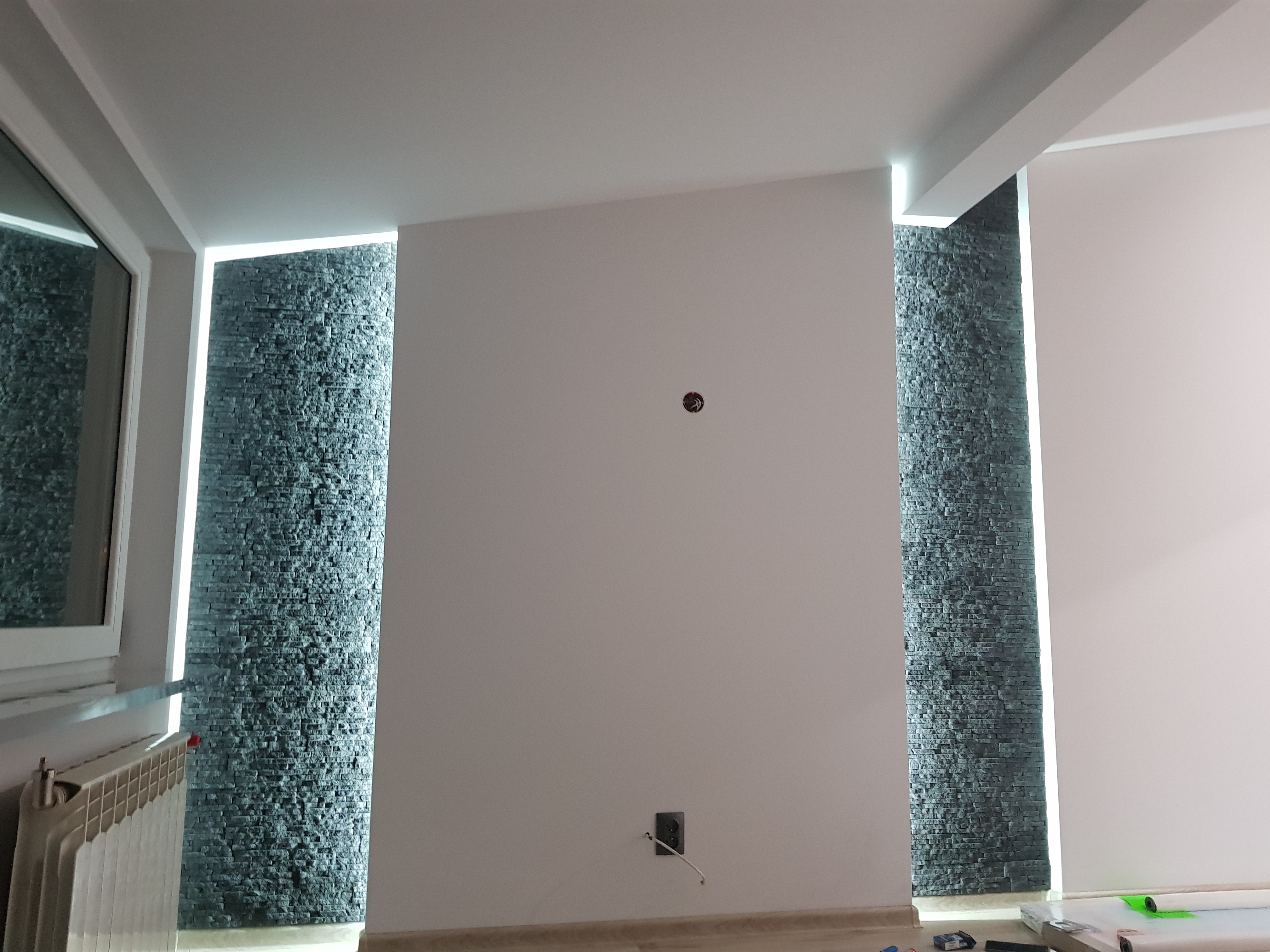 Ściana z dekoracyjną okładziną z kamienia naturalnego, podświetlona pionowymi pasami LED, gniazdko elektryczne na białej ścianie, widoczne narzędzia i materiały budowlane na podłodze.