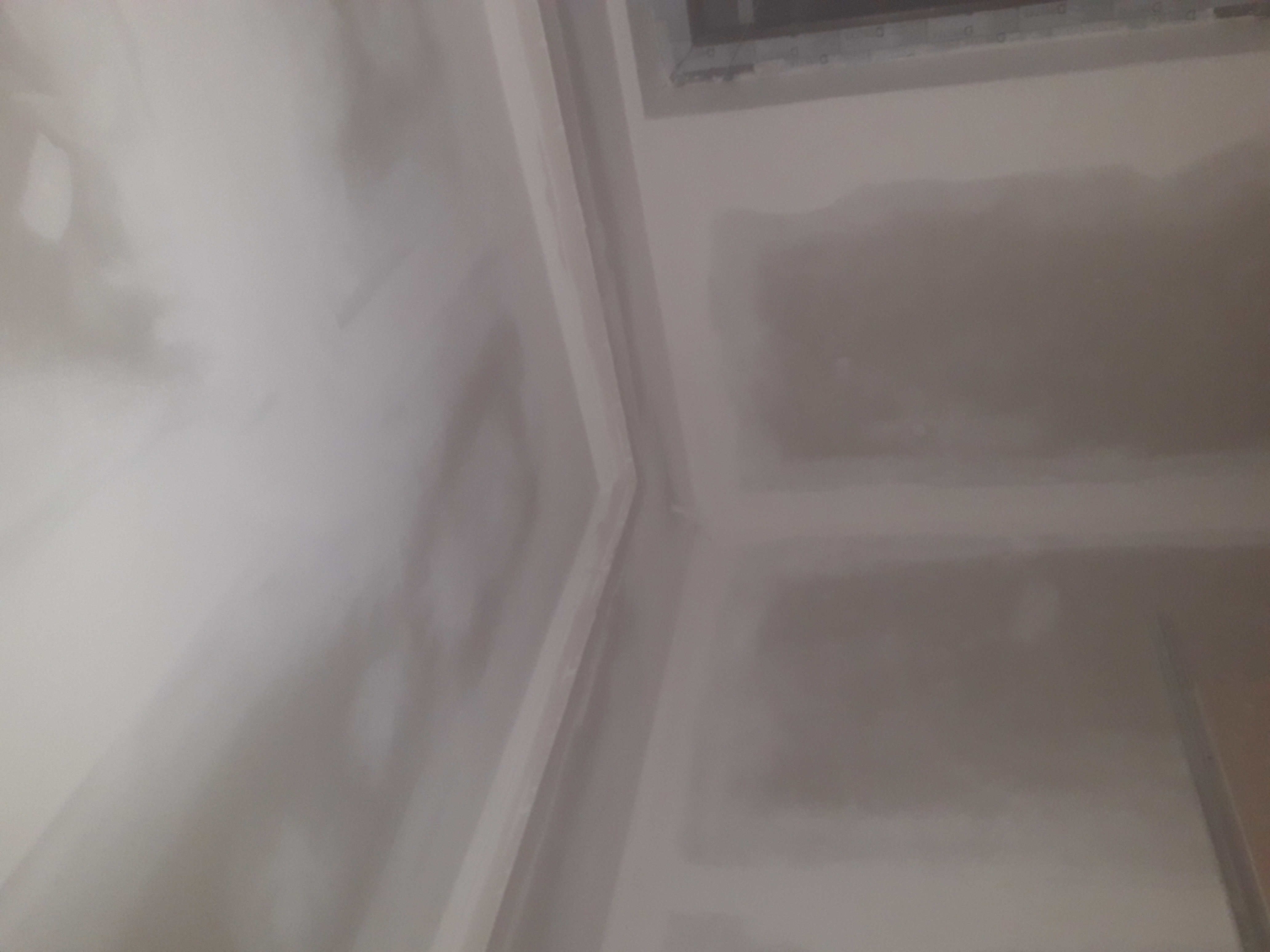 Ściana i sufit w trakcie wygładzania, widoczne nierówności i nałożona warstwa szpachli, okno w górnym rogu.