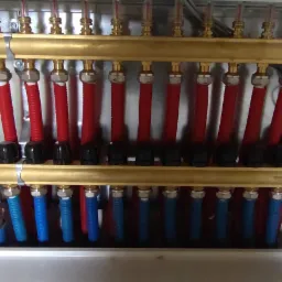 Rozdzielacz ogrzewania podłogowego w metalowej szafce, z czerwonymi i niebieskimi rurami, widoczne zawory i przyłącza.