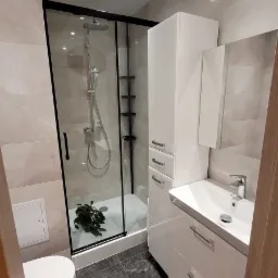 Nowoczesna łazienka z kabiną prysznicową typu walk-in z czarnymi ramami, białą szafką z umywalką i toaletą, wykończona płytkami w odcieniach szarości.