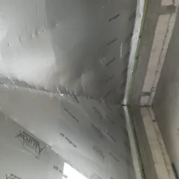 Poddasze z izolacją termiczną Alufox, widoczne zszywki mocujące folię do konstrukcji, ściana z bloczków betonowych.