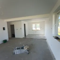 Wnętrze w trakcie remontu: świeżo pomalowane na biało ściany, częściowo położona podłoga, drabina malarska i wiadro z farbą.