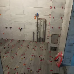 Świeżo ułożone, szare płytki podłogowe i białe ścienne z systemem poziomowania płytek w trakcie prac wykończeniowych w łazience. Widoczne rury instalacyjne i otwory na armaturę.