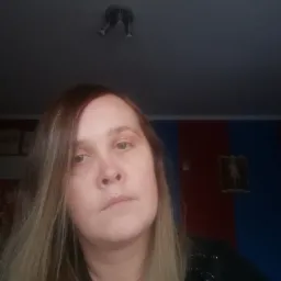 Portret kobiety z długimi blond włosami na tle ściany w kolorach czerwonym i niebieskim, widoczna także lampa sufitowa.