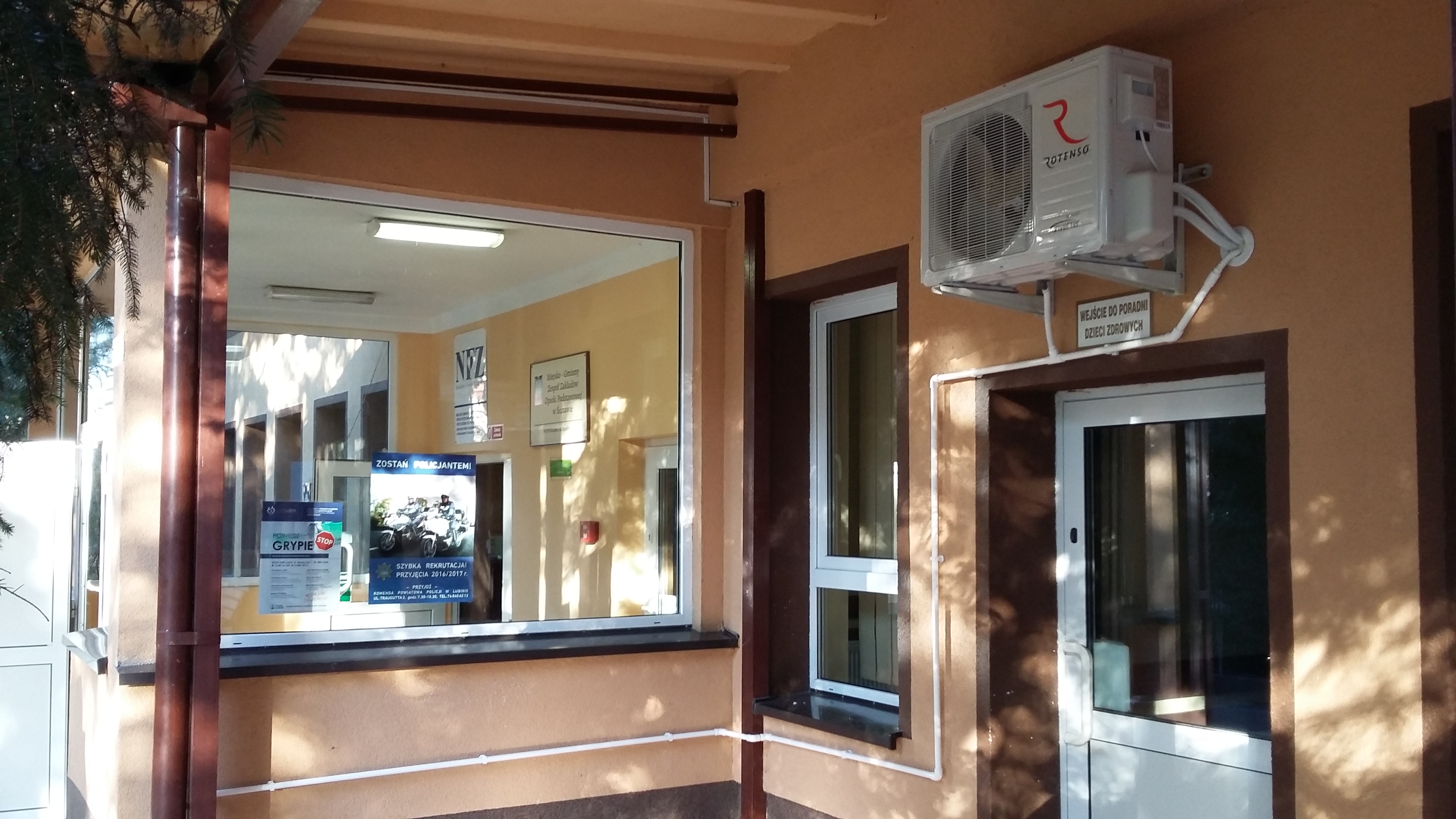 Zewnętrzny klimatyzator Rotenso zamontowany na budynku placówki medycznej, z widocznymi przewodami i tabliczką informacyjną 'Wejście do poradni dzieci zdrowych'.