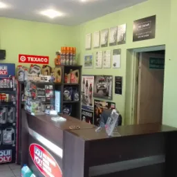 Wnętrze warsztatu samochodowego z ladą recepcyjną, ekspozycją olejów silnikowych Liqui Moly i Texaco, wycieraczek samochodowych oraz certyfikatami na ścianie, widoczny znak ewakuacyjny.