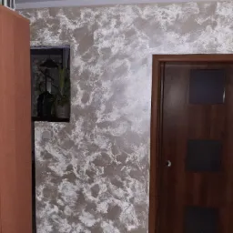 Ściana z dekoracyjną fakturą w odcieniach beżu i bieli, brązowe drzwi z ciemnymi szybkami, fragment szafy z włącznikiem światła oraz widok na roślinę doniczkową w głębi pomieszczenia.