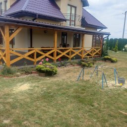 Budujemy z drewna - Dwukondygnacyjny dom z beżową elewacją i ciemnoczerwonym dachem, z drewnianą werandą z ażurowymi balustradami w kolorze jasnego drewna, otoczony ogrodem z niską roślinnością i elementami placu zabaw.