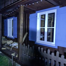 Budujemy z drewna - Fragment drewnianego domu szkieletowego z niebieską elewacją i białymi oknami z okiennicami, widoczne drewno opałowe i girlanda świetlna pod okapem