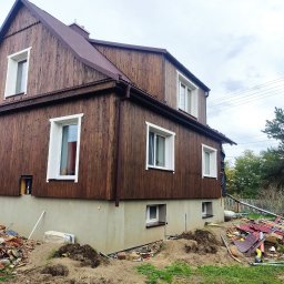 Budujemy z drewna - Wznoszony dom z drewnianą elewacją na betonowym fundamencie, widoczne ślady prac budowlanych i gruz na trawniku.