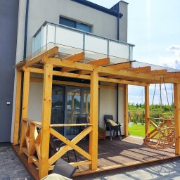 Budujemy z drewna - Nowoczesna pergola z drewna o jasnym odcieniu, z przeszklonym dachem, przylegająca do budynku z balkonem, z wiszącym fotelem typu kokon i drewnianym tarasem.