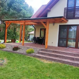 Drewniana pergola z czarnym dachem, przylegająca do domu z beżową elewacją i brązowymi oknami, widok z trawnika na kamienny taras z roślinami ozdobnymi.