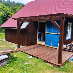 Budujemy z drewna - Dom z czerwonym dachem w trakcie budowy, z drewnianym tarasem i pergolą, widoczne materiały budowlane i narzędzia.