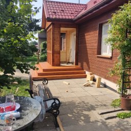Budujemy z drewna - Nowo wybudowany, drewniany ganek z schodami i balustradą, przylegający do brązowego domu z czerwonym dachem, widok z boku z elementami ogrodu i meblami ogrodowymi.