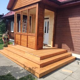 Budujemy z drewna - Nowo wybudowany, drewniany taras z wejściem do domu, świeżo pomalowany na brązowo, z widoczną drabiną wewnątrz i osobą malującą detale na zewnątrz. Widoczna puszka z farbą i narzędzia.