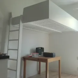 Pok&oacute;j z białym ł&oacute;żkiem na antresoli z drabinką, pod spodem drewniany st&oacute;ł z narzędziami w pudełkach, oświetlony lampą sufitową.