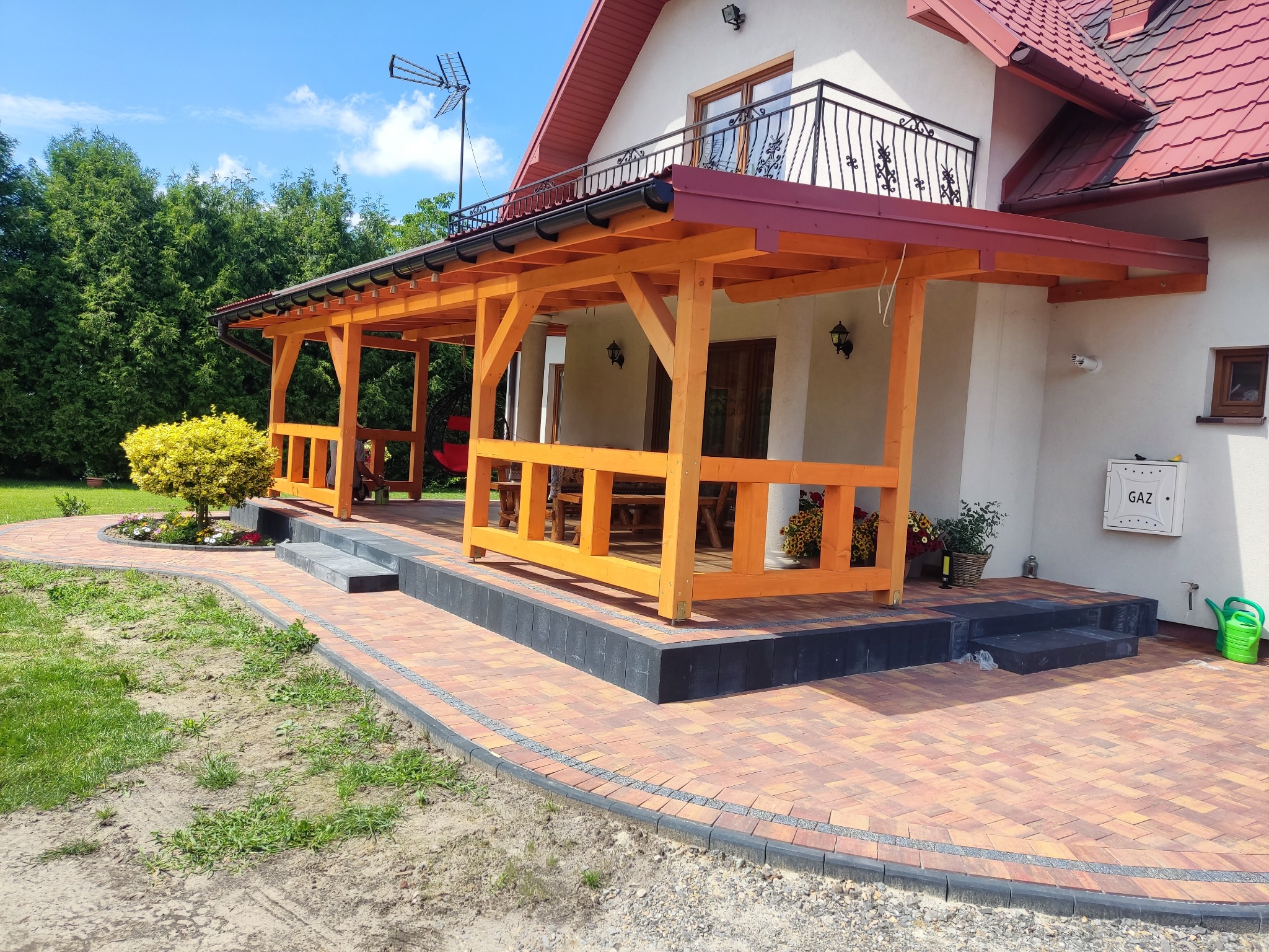 Zadaszony taras z surowego drewna, połączony z domem, z widocznymi schodami wejściowymi i kostką brukową na podwórku.