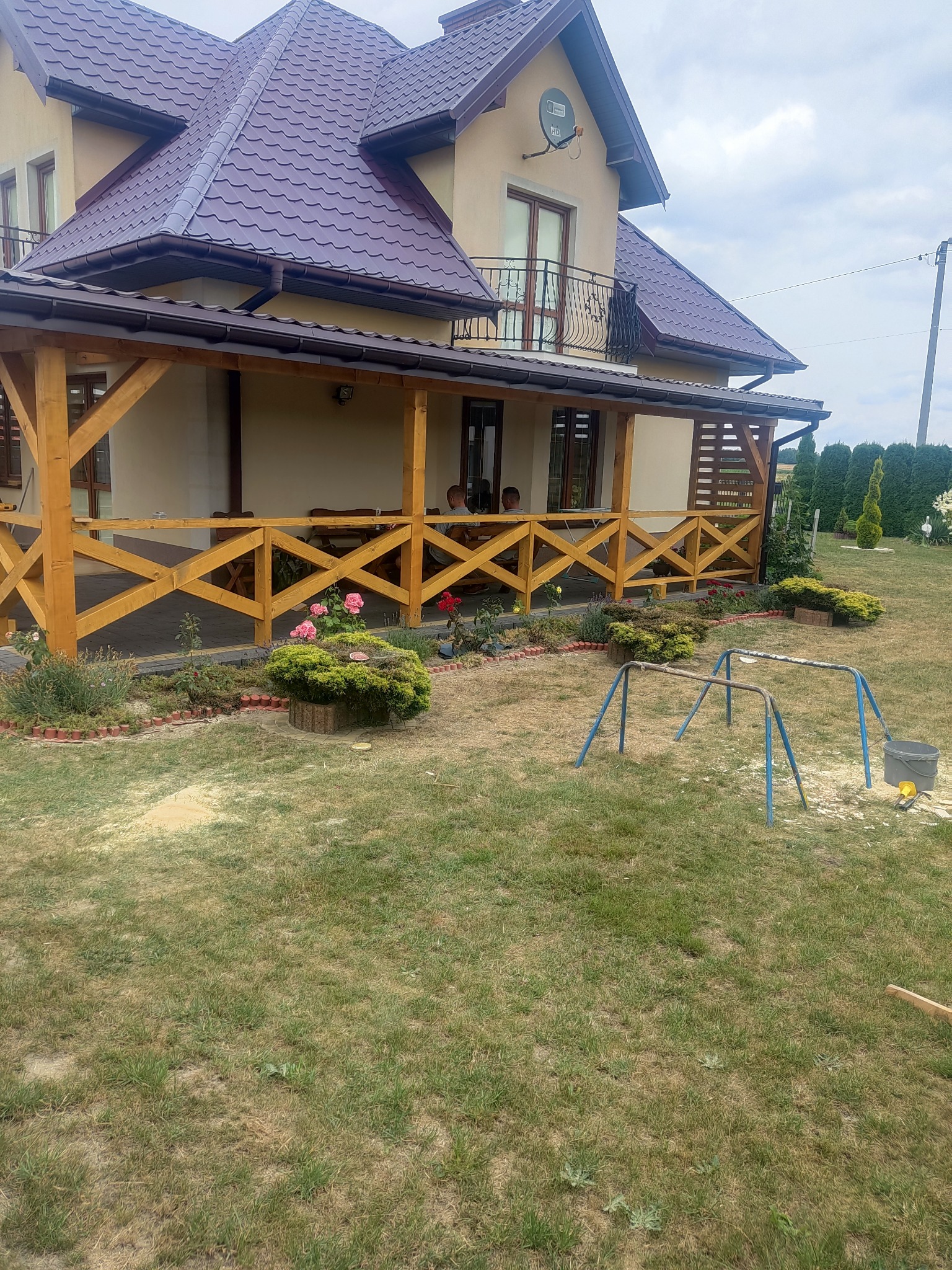 Dwukondygnacyjny dom z beżową elewacją i ciemnoczerwonym dachem, z drewnianą werandą z ażurowymi balustradami w kolorze jasnego drewna, otoczony ogrodem z niską roślinnością i elementami placu zabaw.