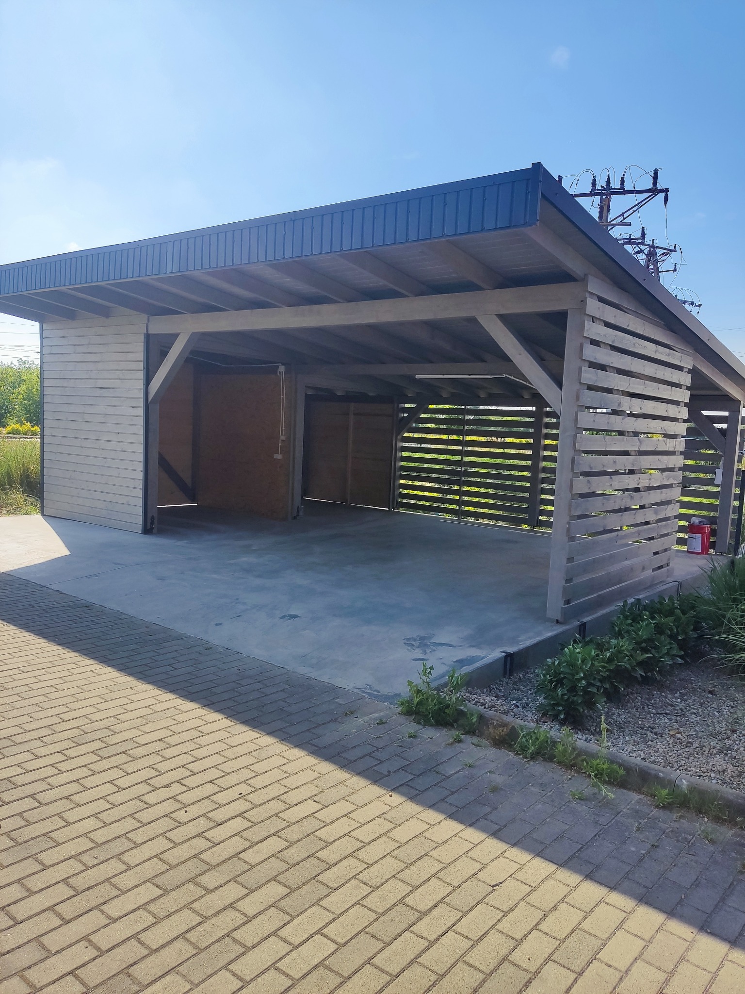 Nowoczesna, otwarta wiata garażowa z drewnianą konstrukcją i betonową posadzką, z widocznymi elementami oświetlenia i roślinnością w tle, sfotografowana w słoneczny dzień.