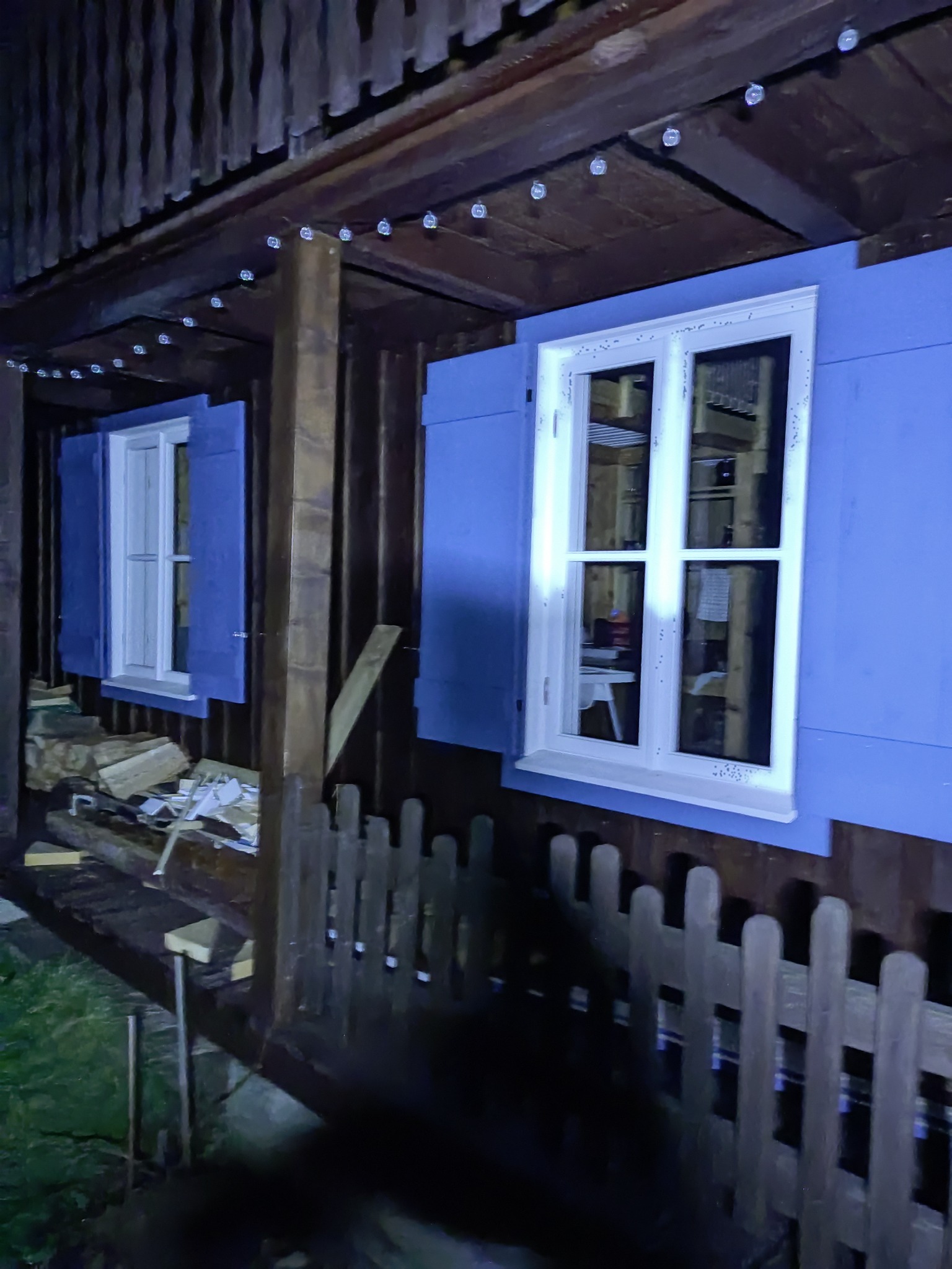 Fragment drewnianego domu szkieletowego z niebieską elewacją i białymi oknami z okiennicami, widoczne drewno opałowe i girlanda świetlna pod okapem