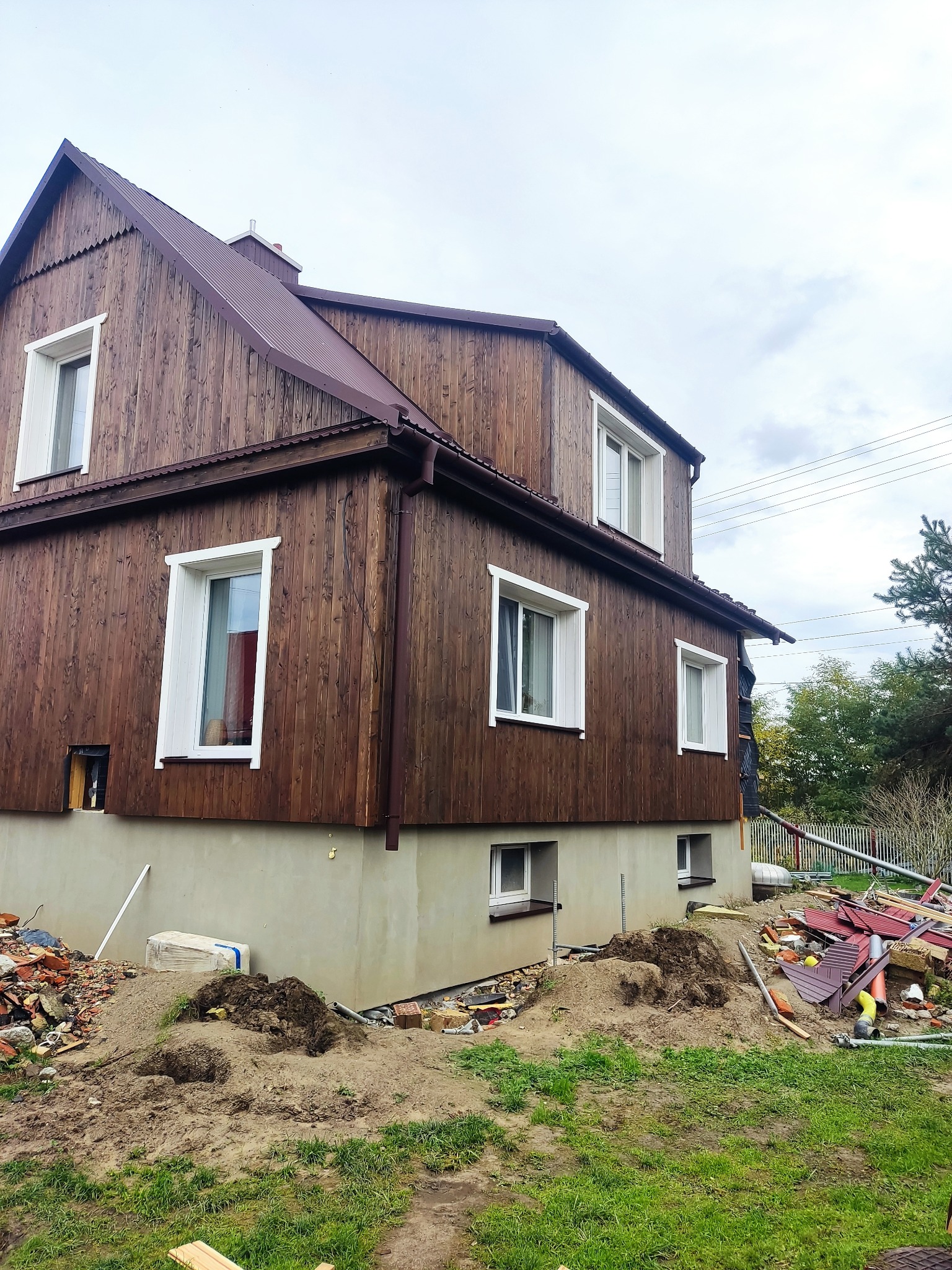 Wznoszony dom z drewnianą elewacją na betonowym fundamencie, widoczne ślady prac budowlanych i gruz na trawniku.