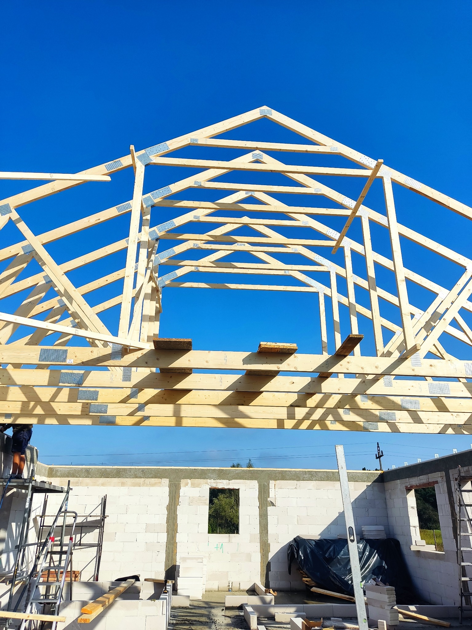 Konstrukcja drewniana dachu w trakcie budowy domu z bloczków, widok na niebieskie niebo, drabina rusztowania i poziomica przy ścianie.