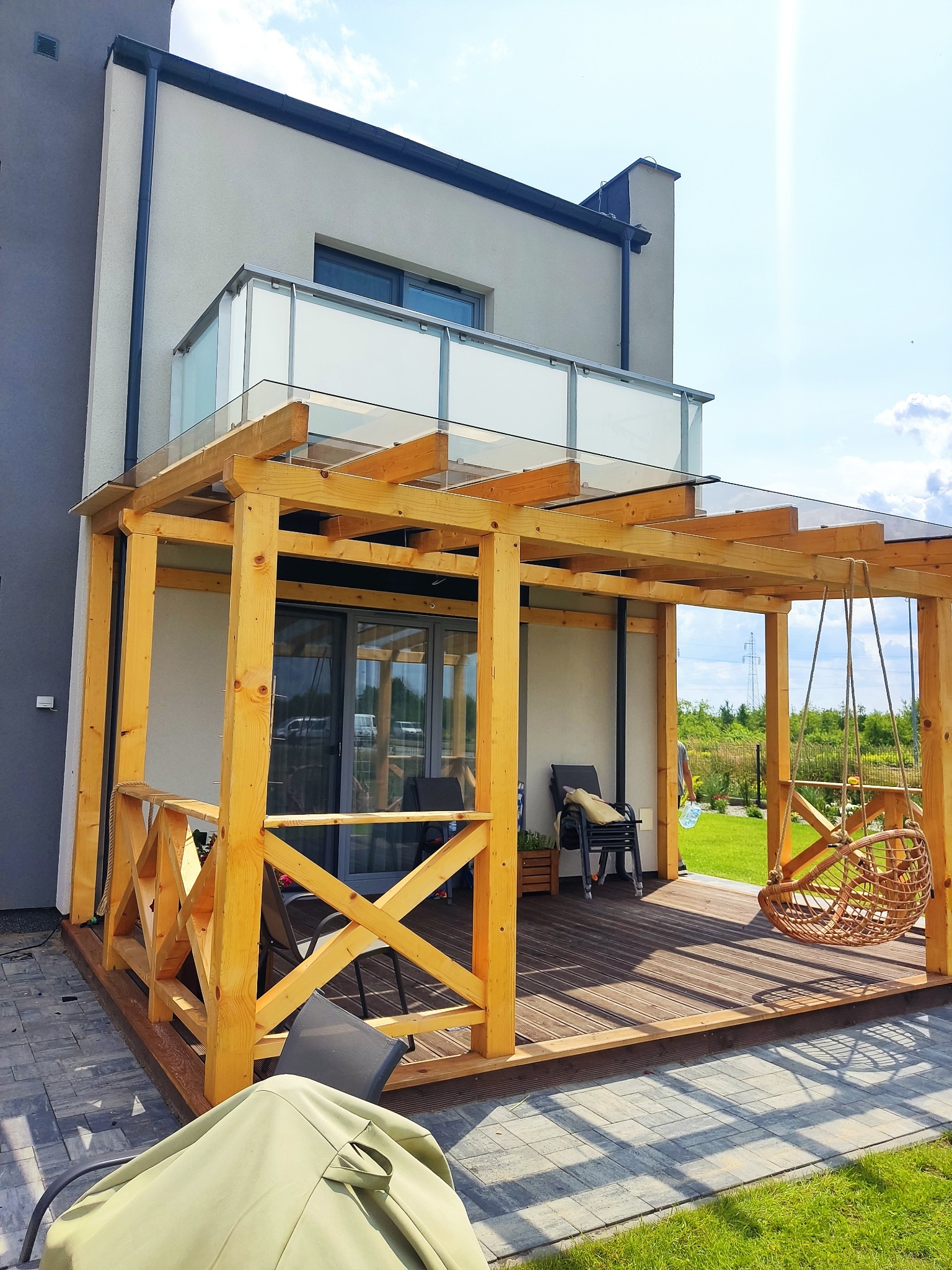 Nowoczesna pergola z drewna o jasnym odcieniu, z przeszklonym dachem, przylegająca do budynku z balkonem, z wiszącym fotelem typu kokon i drewnianym tarasem.
