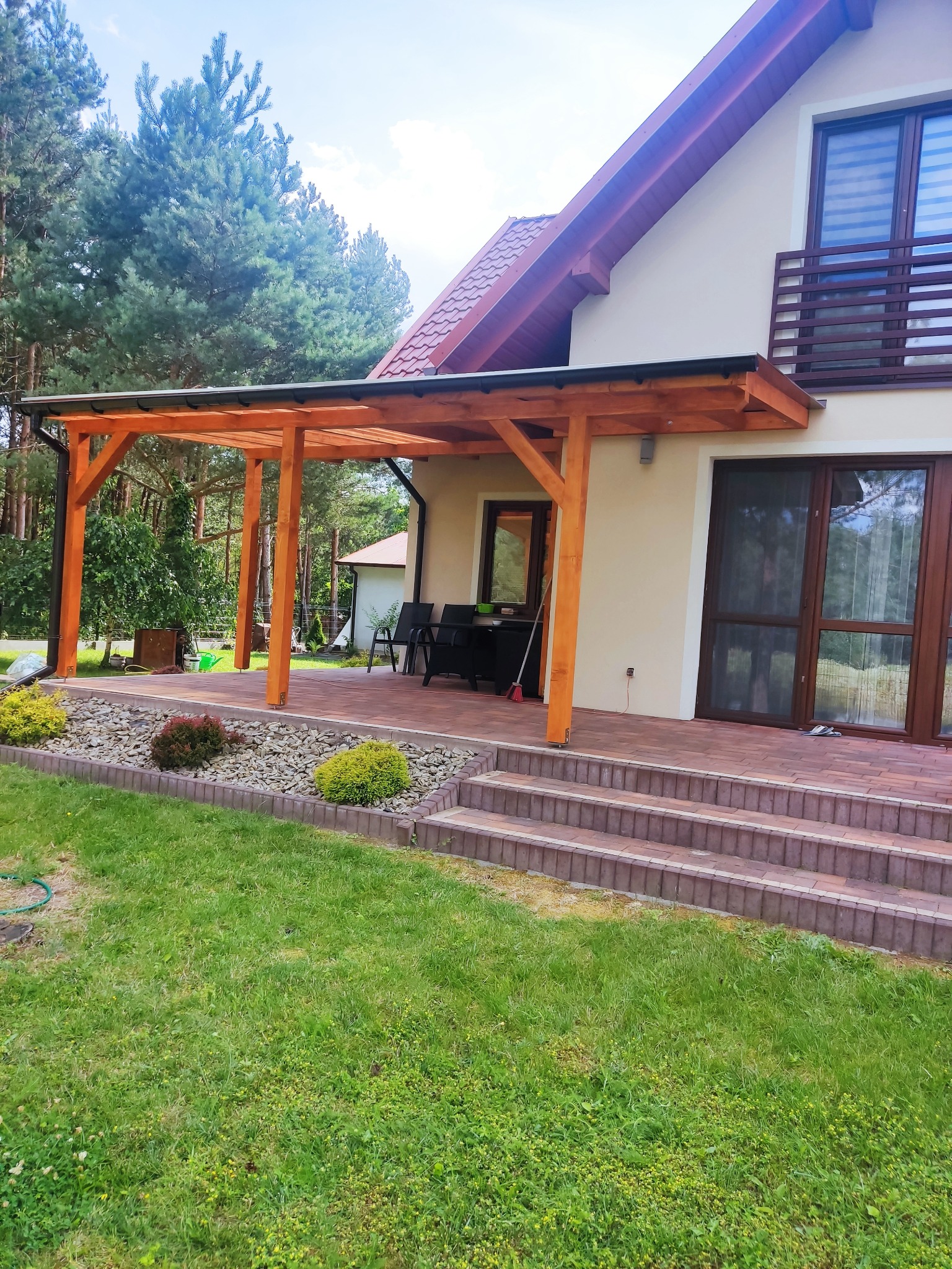 Drewniana pergola z czarnym dachem, przylegająca do domu z beżową elewacją i brązowymi oknami, widok z trawnika na kamienny taras z roślinami ozdobnymi.