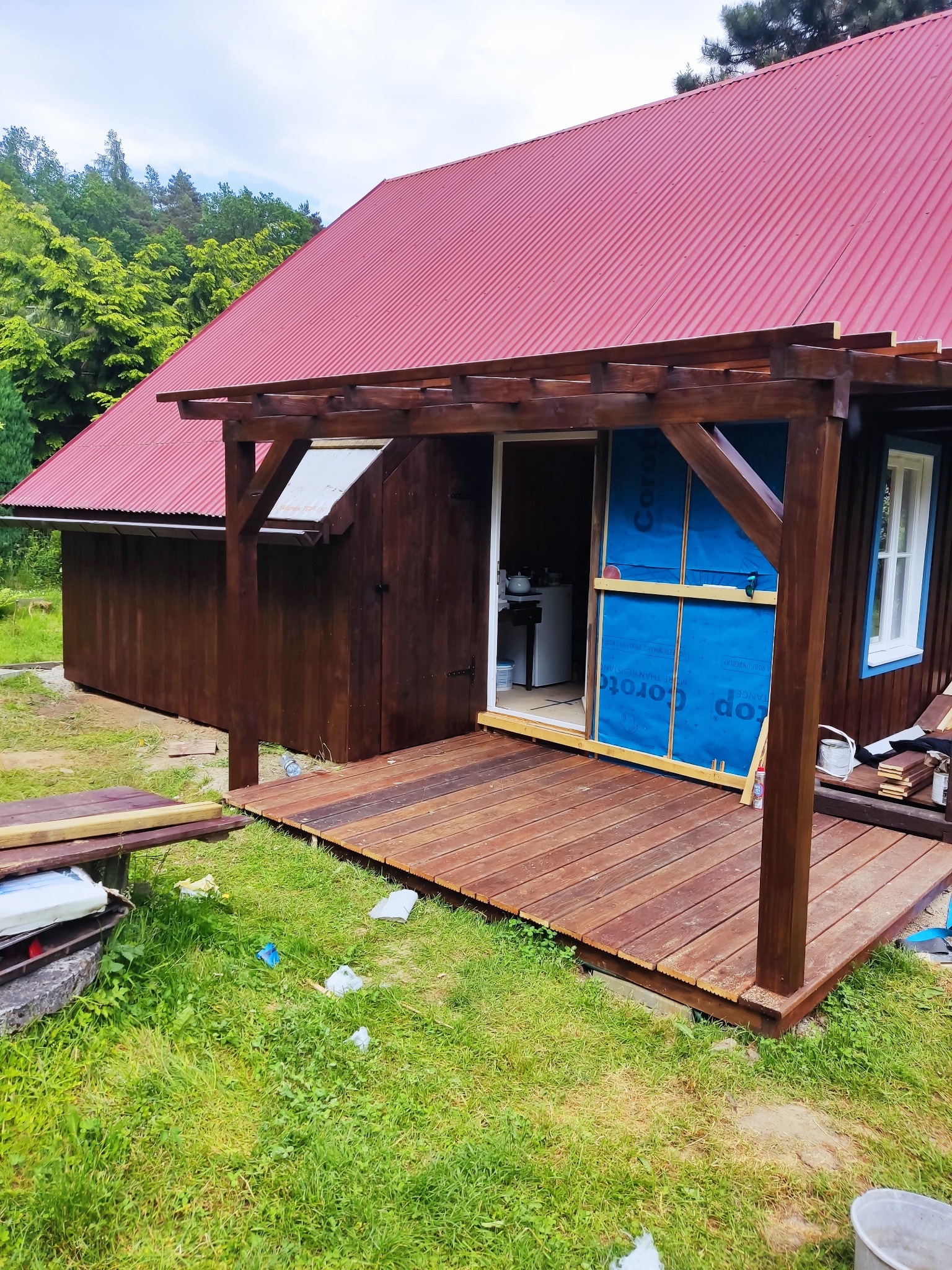 Dom z czerwonym dachem w trakcie budowy, z drewnianym tarasem i pergolą, widoczne materiały budowlane i narzędzia.